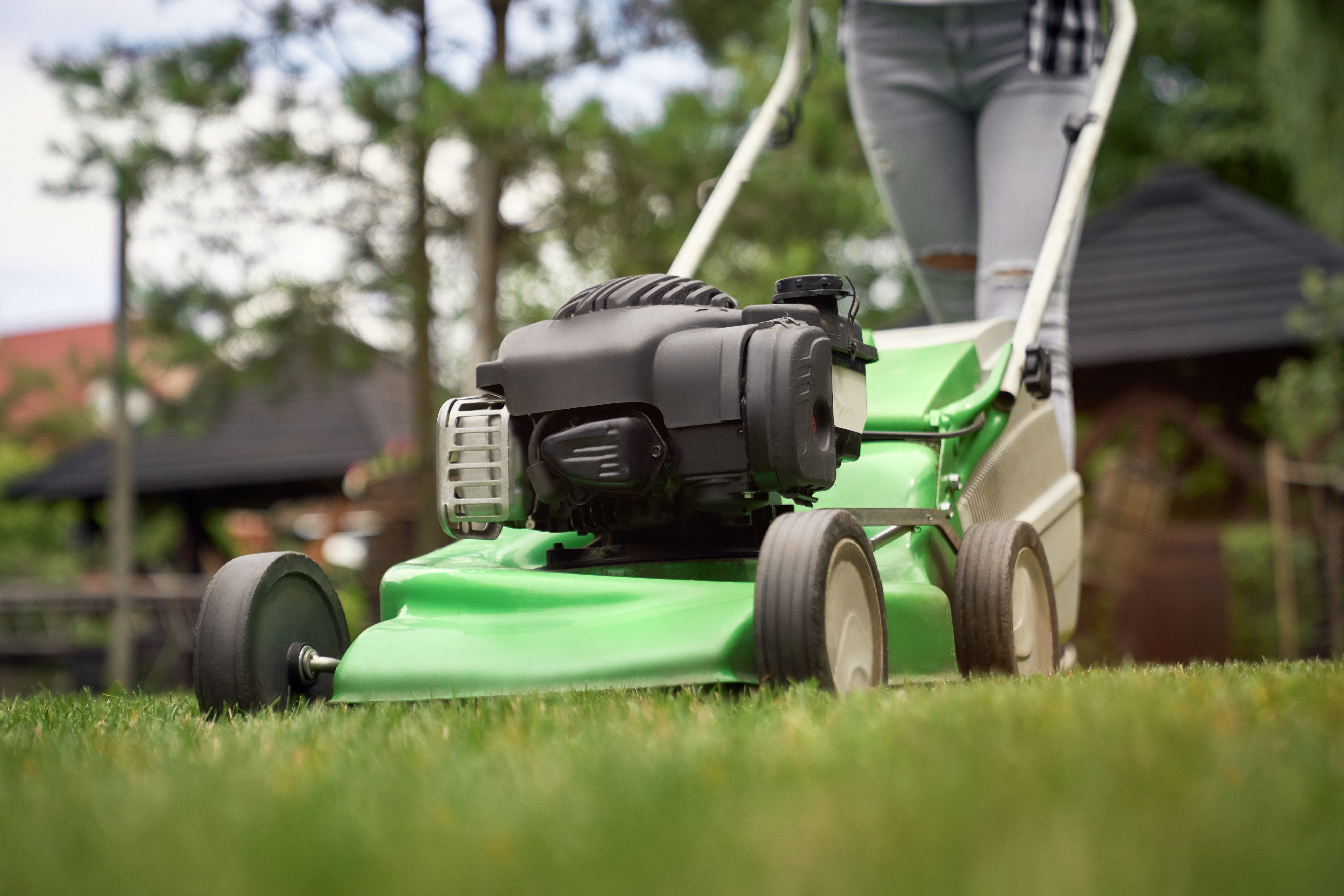 Close up of female legs of gardener using green lawn mower on backyard. Selective focus of woman working in summer, cutting grass in backyard. Concept of gardening, work, nature.